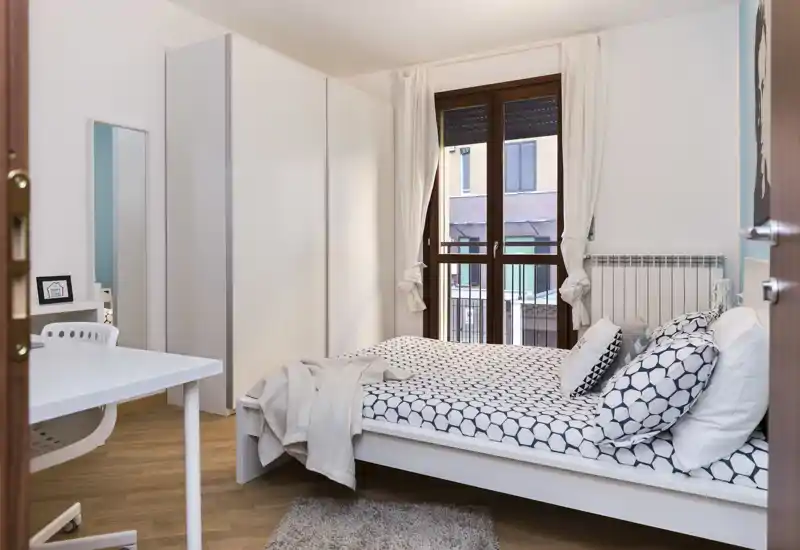 Dormitorio moderno y luminoso con cama doble con ropa de cama estampada, gran armario y escritorio junto a la ventana que ofrece luz natural y vistas a la calle.