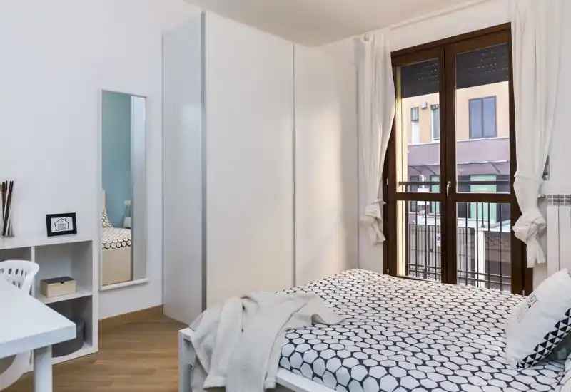 Dormitorio moderno y luminoso con cama doble bien hecha con funda de patrón geométrico, amplio armario blanco y ventana con vista a la calle.