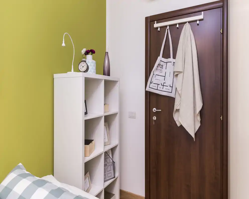 Angolo di camera da letto con vista parziale del letto, un mobiletto bianco a cubi e una porta in legno con appendini. Lo spazio è ordinato e semplicemente arredato con tessuti morbidi e toni pastello.