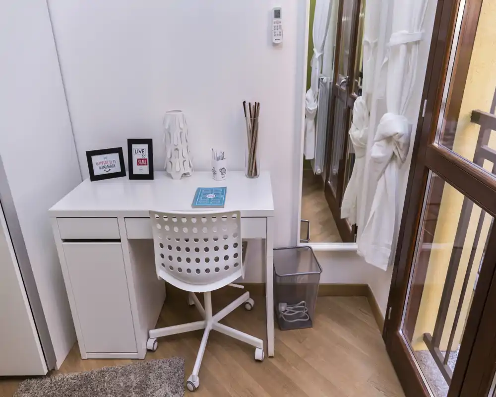 Angolo living/working luminoso e compatto con scrivania bianca, sedia girevole traforata e porta balcone che offre luce naturale.