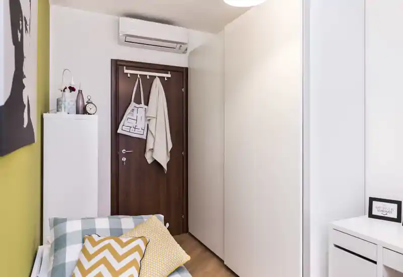 Angolo di camera da letto piccolo e ben curato con cuscini decorativi, biancheria e un ampio armadio bianco di fronte a una porta in legno; luminoso, moderno e accogliente.