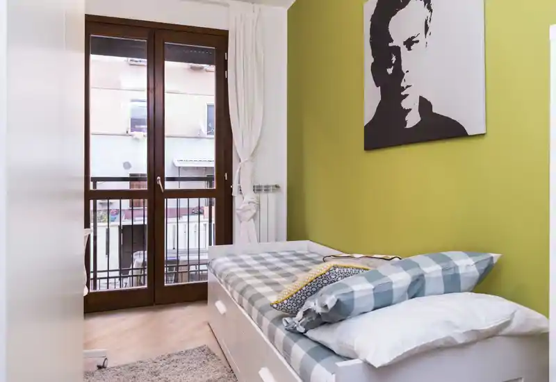Camera singola arredata con letto appoggiato al muro, porta finestra verso il balcone e quadro moderno su una parete verde. Lenzuola pulite, cuscini decorativi e un tappeto morbido rendono l'ambiente accogliente e luminoso, ideale per affitti brevi.