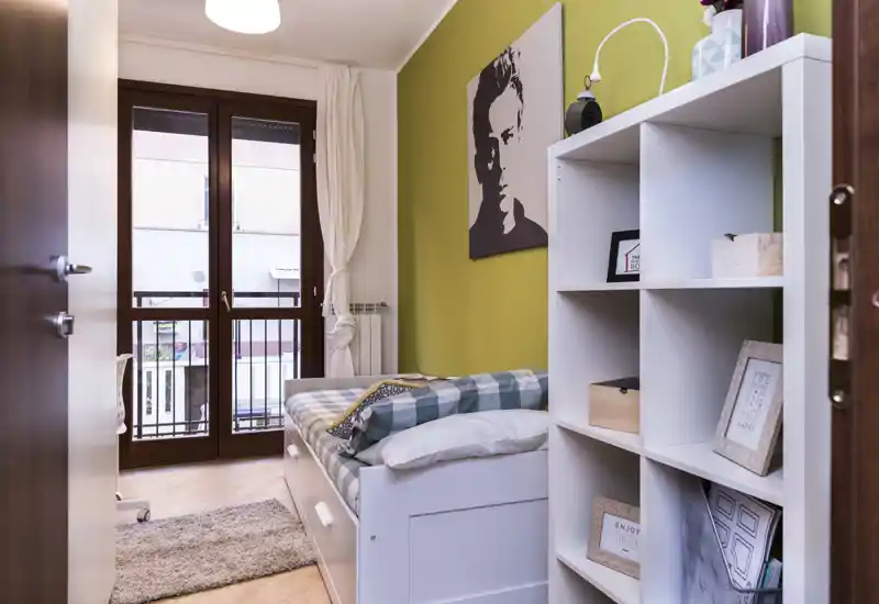 Camera singola compatta e stilosa con letto/giorno, biancheria a quadri, libreria a cubi e ampia finestra che garantisce luce naturale.