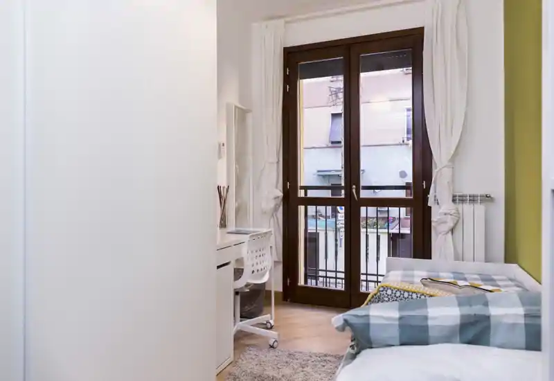 Camera luminosa e accogliente con letto singolo vicino a una grande porta finestra, tessili morbidi e una piccola scrivania — ideale per un soggiorno confortevole in città.