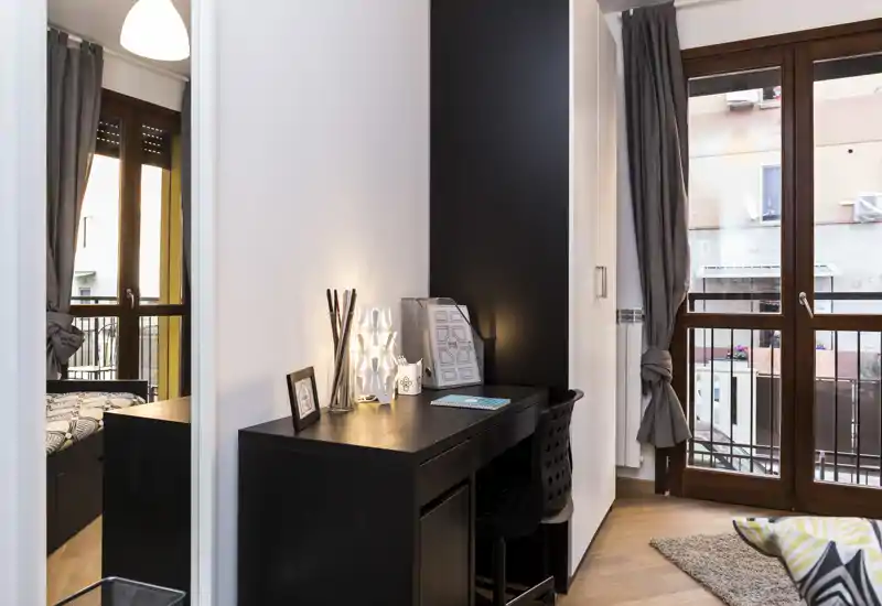 Rincón de dormitorio luminoso y moderno con escritorio negro, lámpara decorativa y vista parcial de la cama junto a la ventana — perfecto para un rincón de trabajo acogedor.