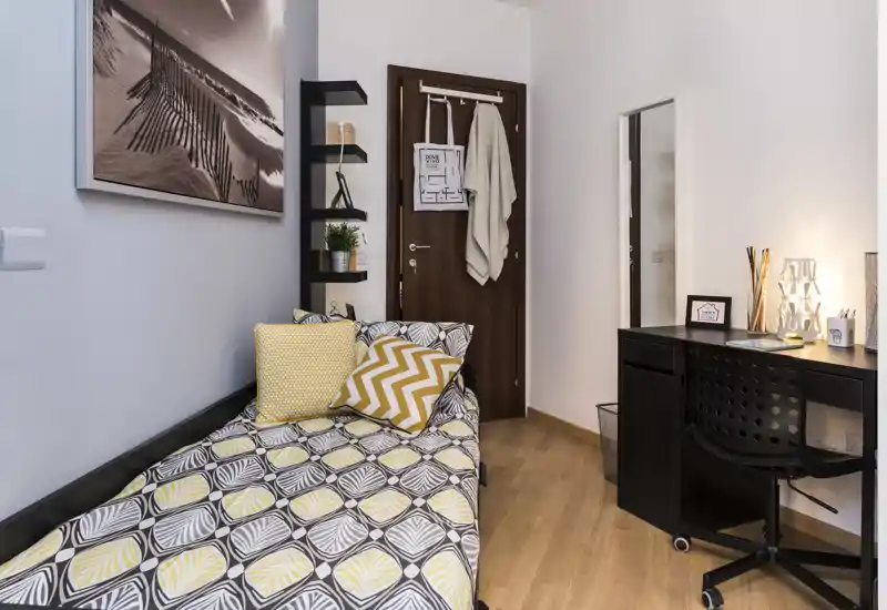 Dormitorio pequeño y bien presentado con cama individual, ropa de cama estampada y cojines coloridos, escritorio compacto con lámpara y silla, suelo de madera cálida — luminoso y acogedor para una persona.