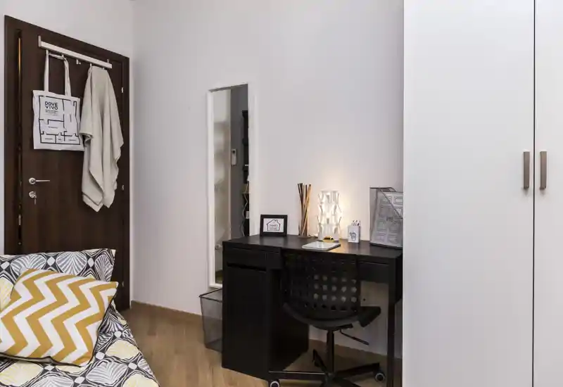 Angolo camera accogliente e ben arredato con letto a fantasia, scrivania e armadio; la luce calda della lampada crea un angolo lavoro invitante accanto al letto.