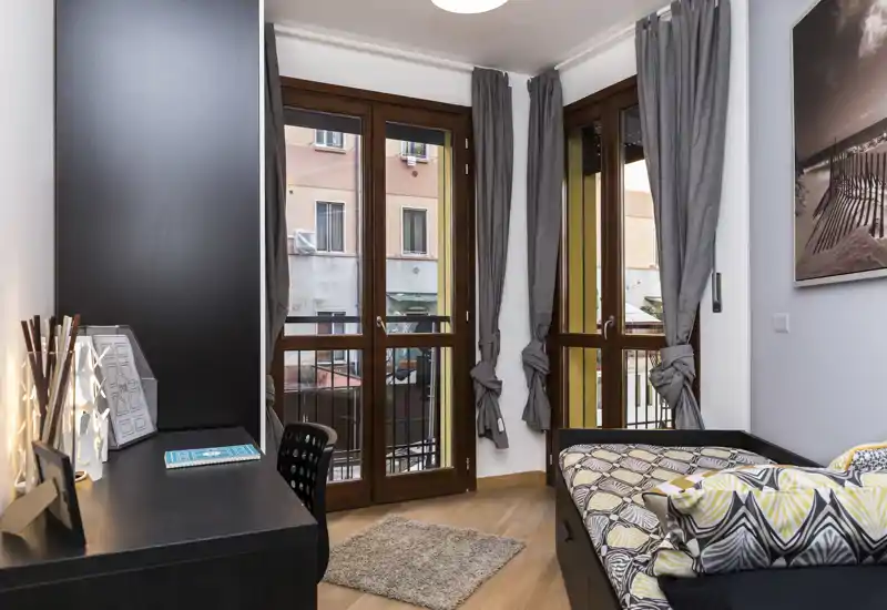 Dormitorio compacto con cama individual, cojines estampados y una alfombra pequeña; grandes ventanas con marco de madera aportan luz natural y cortinas grises de estilo moderno.