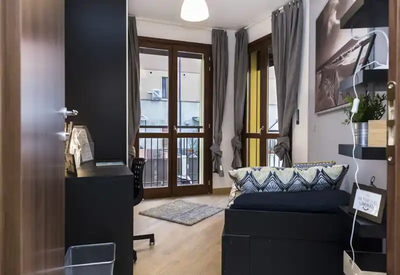 Dormitorio moderno y luminoso con cama individual, cojines decorativos y grandes ventanas que permiten mucha luz natural.