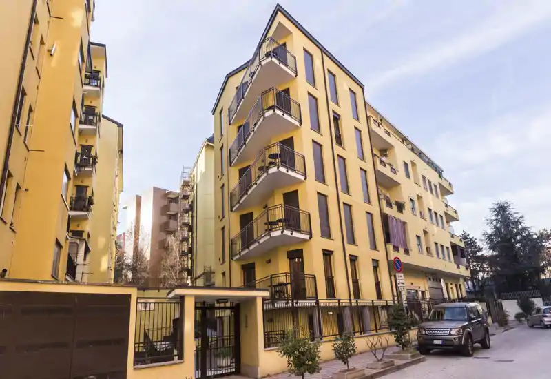 Bright exterior view of a yellow residential building with multiple balconies and a gated entrance, photographed from the street under clear daylight.