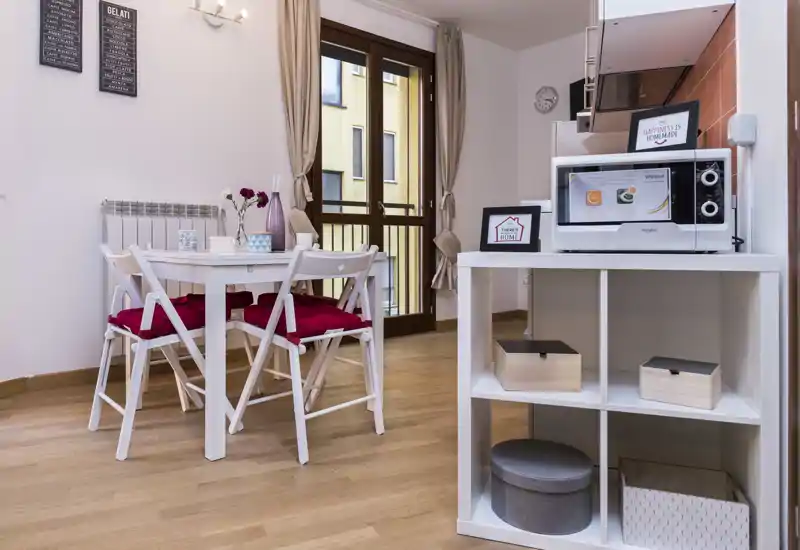 Bright, tidy dining nook with a small table and four chairs, red cushions, and a balcony door bringing natural light. A white shelving unit with a microwave and decorative boxes frames the space.