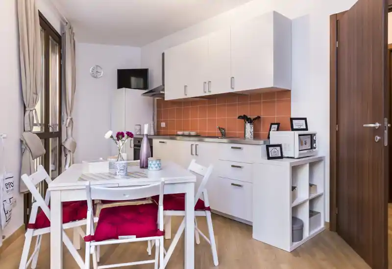 Compact, fully equipped kitchen with white cabinets, orange tiled backsplash and a cozy dining table set with red cushions — bright and neatly staged for short-term rental photos.