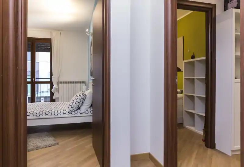 Bright, neatly styled bedroom seen through a doorway: single/double bed with patterned linens, pillows and a small rug near a window providing natural light.