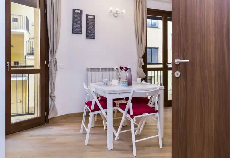 Bright, modern dining area with a small white table and four folding chairs dressed with red cushions, set near large windows that provide natural light.