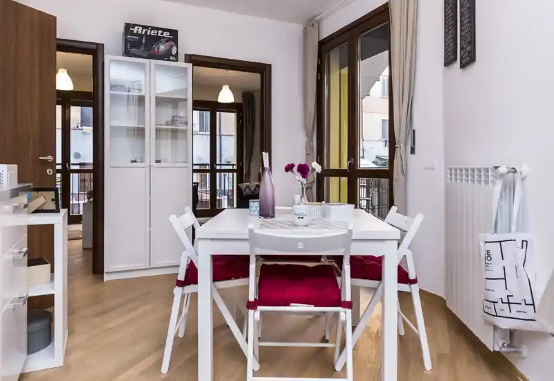 Bright, modern dining area with a white table, four chairs dressed with red cushions and decorative accents like a vase and placemat; large windows provide natural light and a pleasant atmosphere.
