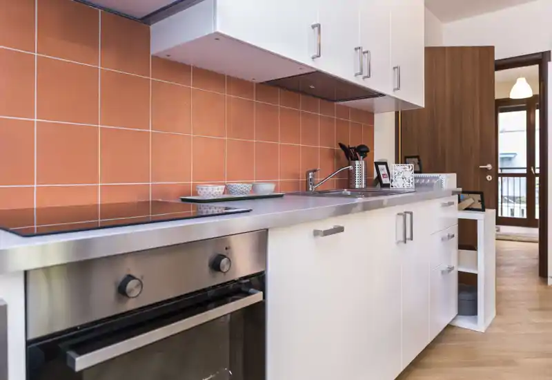 Bright modern kitchen with white cabinets, stainless-steel countertop and orange tiled backsplash; built-in oven and induction hob visible, plus sink and utensils for a practical cooking space.