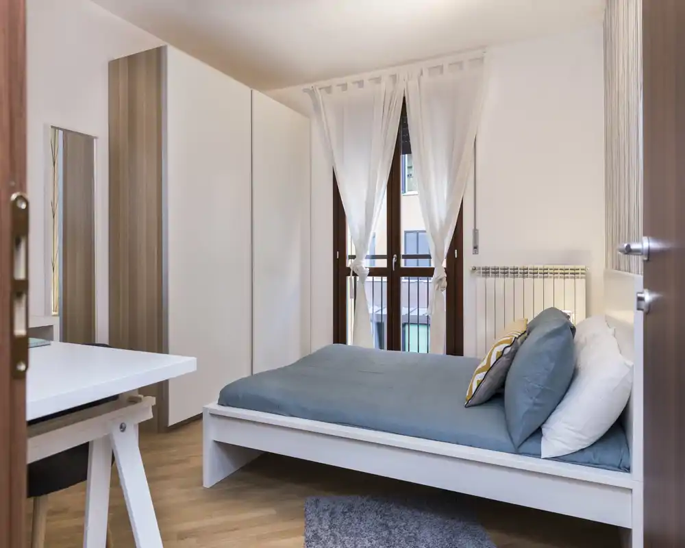 Camera da letto luminosa e ben allestita con letto matrimoniale, biancheria blu e cuscini decorativi; armadio e scrivania visibili, finestra con tende leggere che lasciano entrare luce naturale.