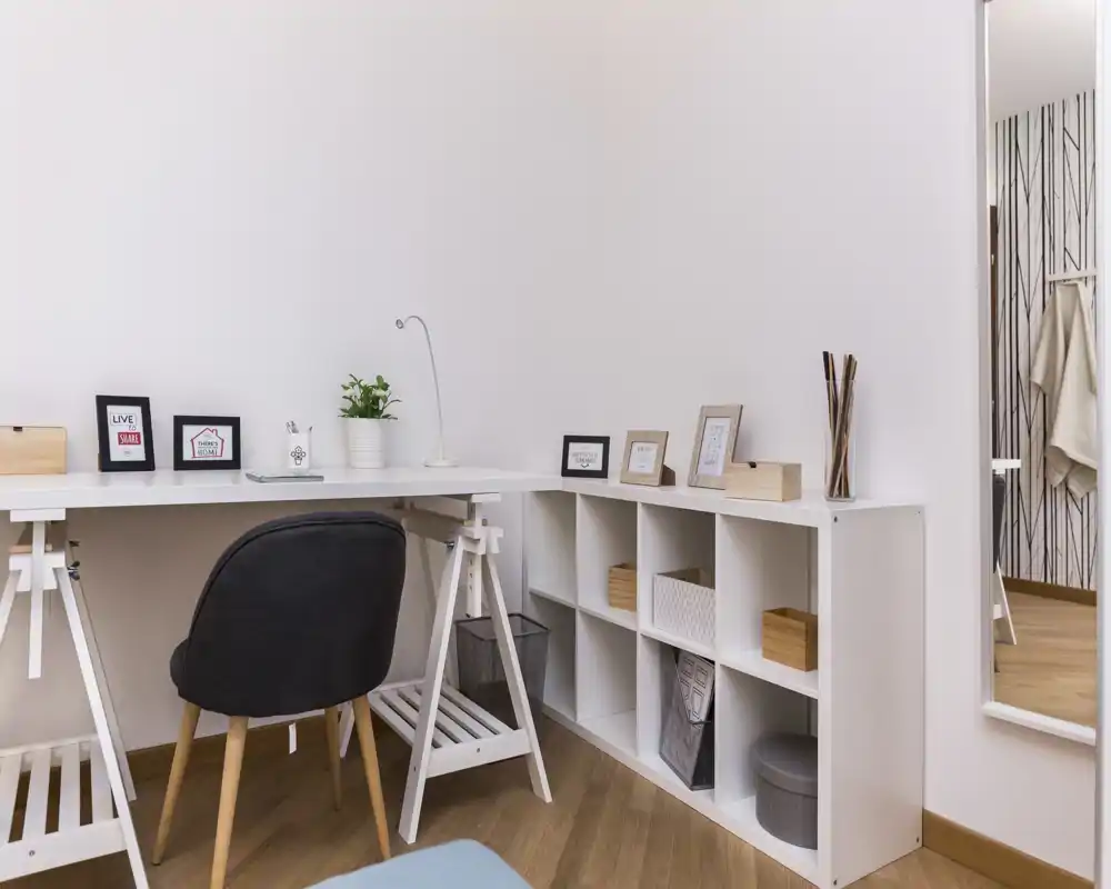 Angolo living/studio compatto e moderno con scrivania bianca, sedia nera e libreria bassa; toni neutri e piccoli oggetti decorativi creano un ambiente funzionale e rilassante.