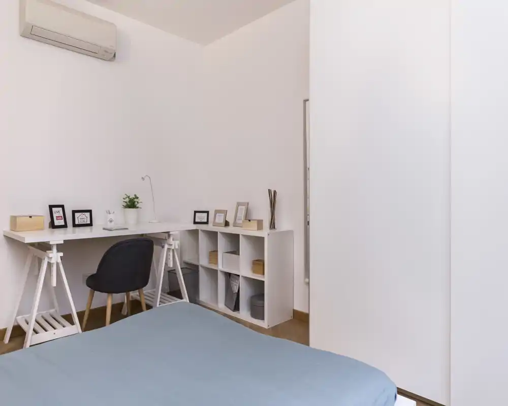 Camera da letto minimalista con letto ordinato in primo piano, una scrivania bianca e un mobile a cubi; luce naturale tenue che crea un'atmosfera rilassante.