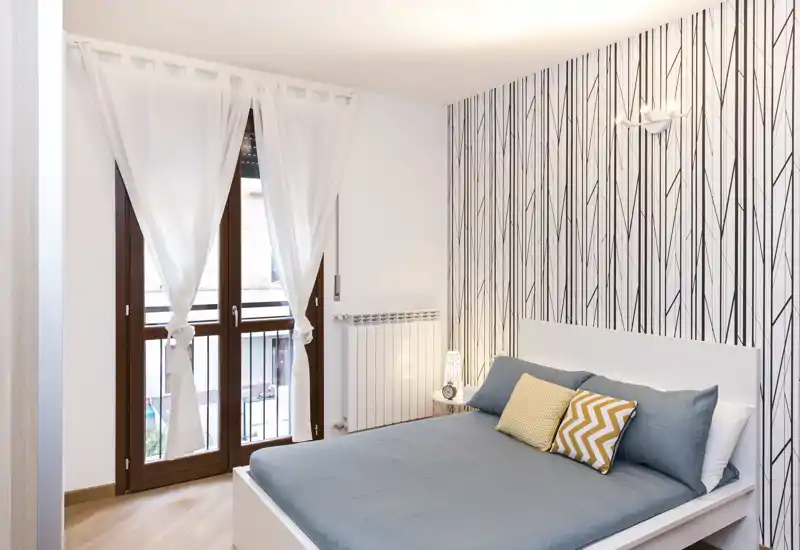 Camera da letto moderna e luminosa con letto matrimoniale in lenzuola grigie, cuscini decorativi gialli e una parete con carta da parati a motivo; ampia finestra con tende leggere che garantisce molta luce naturale.