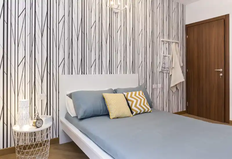 Camera da letto moderna e luminosa con testiera bianca, biancheria blu e cuscini gialli decorativi; carta da parati a motivo e lampada da comodino creano un'atmosfera accogliente ed elegante.
