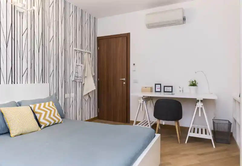 Camera da letto moderna e luminosa con un grande letto coperto da lenzuola blu, cuscini decorativi e una piccola zona studio con scrivania bianca e sedia; carta da parati a righe e pavimento in legno caldo.