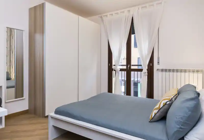 Camera da letto moderna e luminosa con letto matrimoniale, cuscini decorativi e un ampio armadio; luce naturale dalle finestre alte e stile contemporaneo semplice.