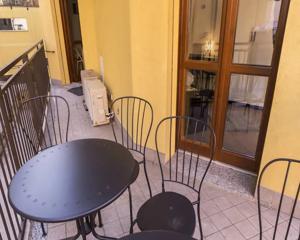 Balcone soleggiato e stretto con tavolo rotondo in metallo e sedie abbinate, pavimento piastrellato e porta a vetri che conduce nell'appartamento — ideale per un caffè mattutino.
