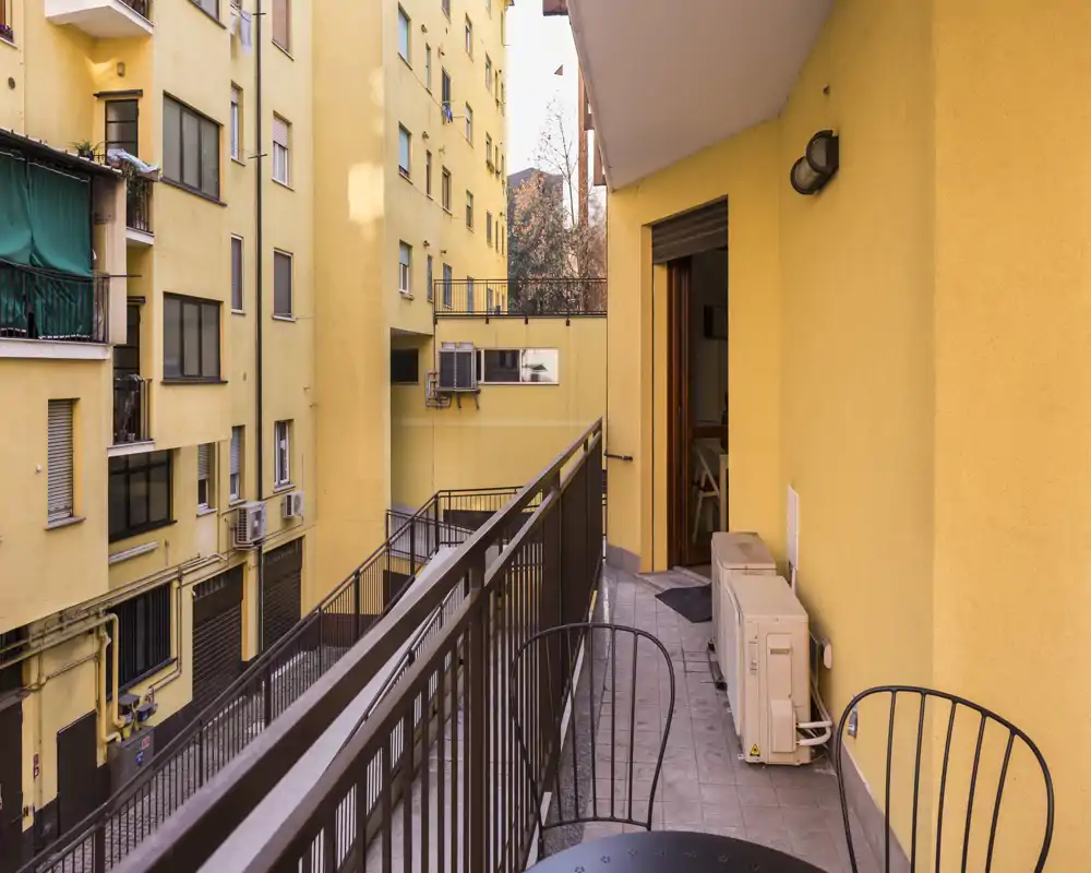 Balcone stretto e soleggiato con ringhiera in metallo, piccolo tavolo e sedie e unità di condizionamento esterne; ideale per un caffè mattutino in un contesto urbano centrale.