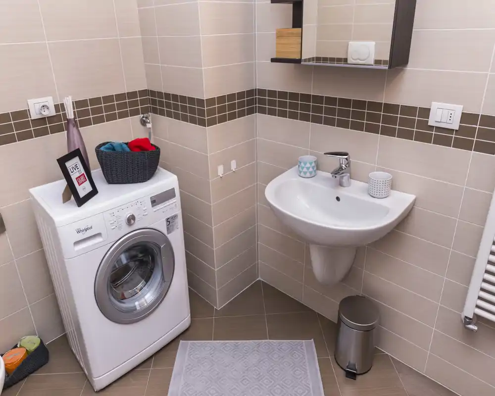 Bagno compatto con lavabo a parete, piastrelle e lavatrice, ben organizzato e luminoso.