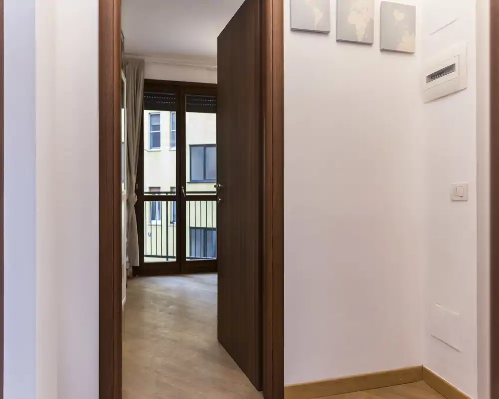 Piccolo ingresso/corridoio pulito che conduce a una stanza interna con porta in legno e pavimento in parquet; pareti chiare e luce naturale proveniente dalla stanza oltre.