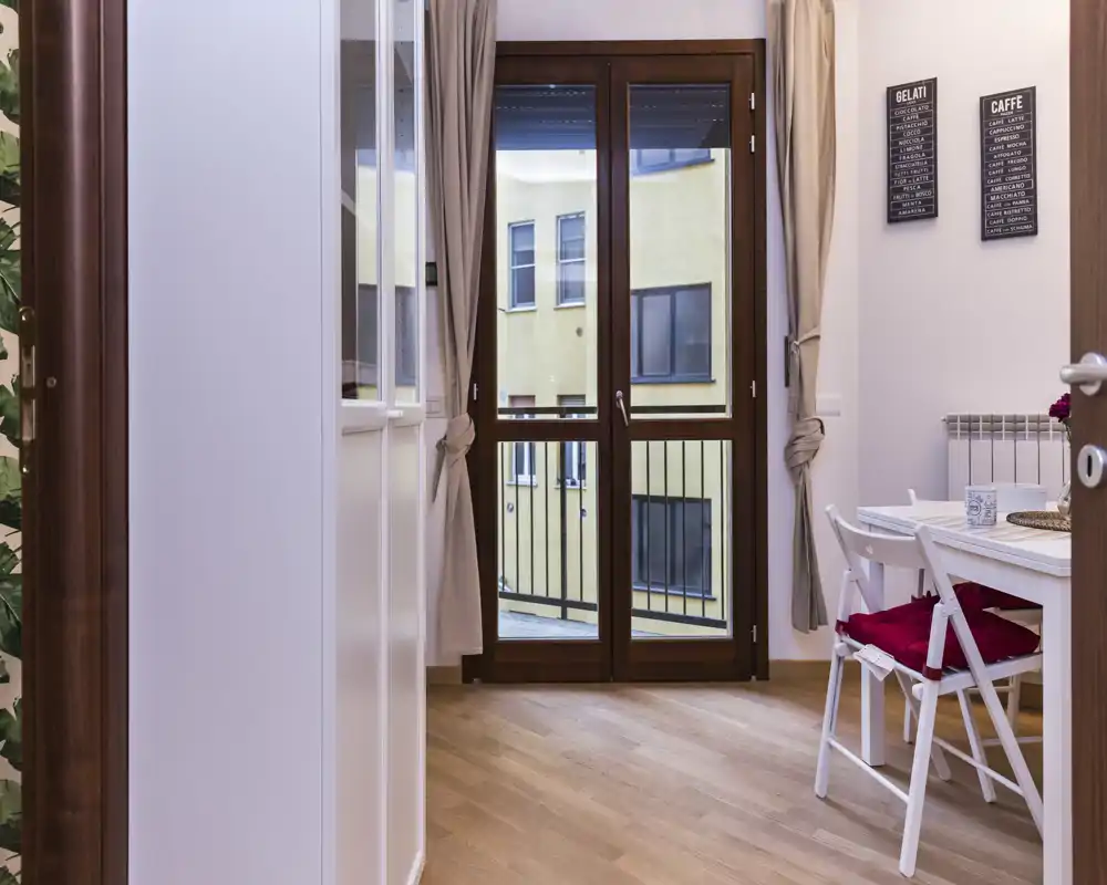 Accogliente angolo pranzo ben allestito con un piccolo tavolo e sedie pieghevoli vicino alla porta del balcone; la luce naturale e il pavimento in legno caldo creano un ambiente invitante.
