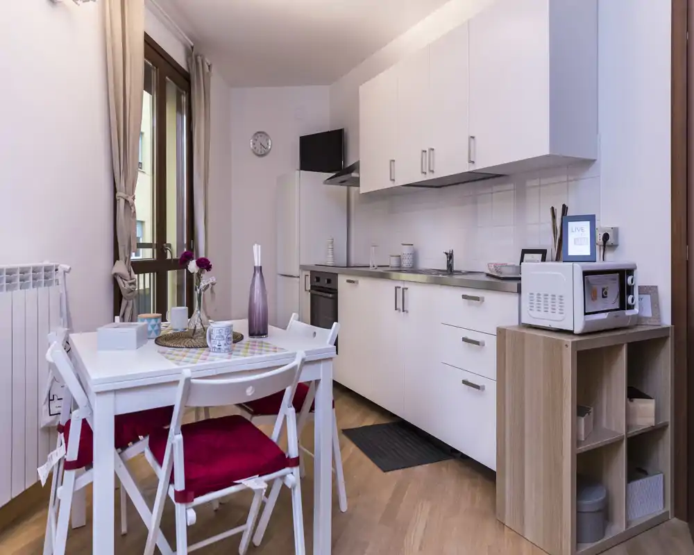 Cucina compatta e moderna con ante bianche, forno incassato, frigorifero e piccolo tavolo da pranzo con cuscini rossi — luminosa, ordinata e accogliente per pasti rilassati.