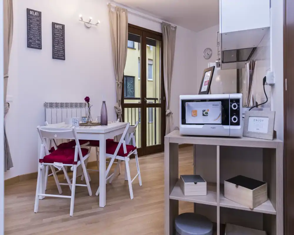Zona pranzo luminosa e compatta con un tavolo bianco e quattro sedie con cuscini rossi, vicino a una porta finestra che garantisce luce naturale. Stile moderno e semplice con dettagli decorativi, ideale per appartamenti piccoli.