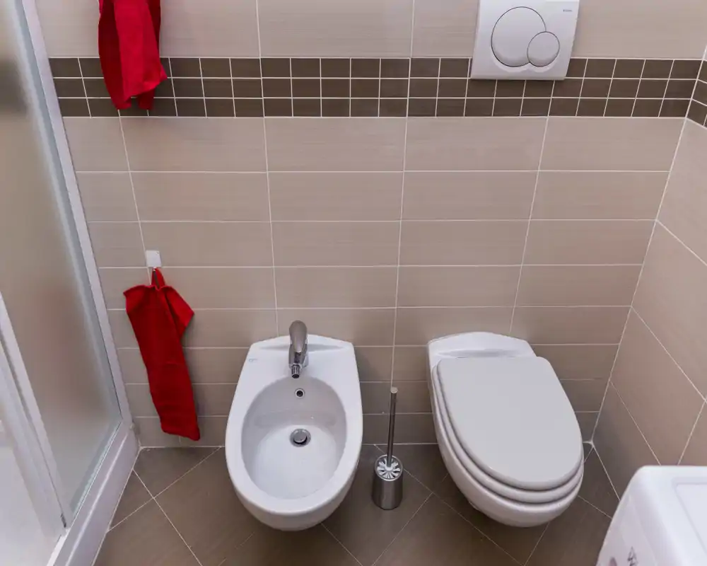 Angolo di bagno moderno e pulito con bidet e WC chiuso su pareti e pavimento piastrellati beige, con asciugamani rossi che danno un tocco di colore.