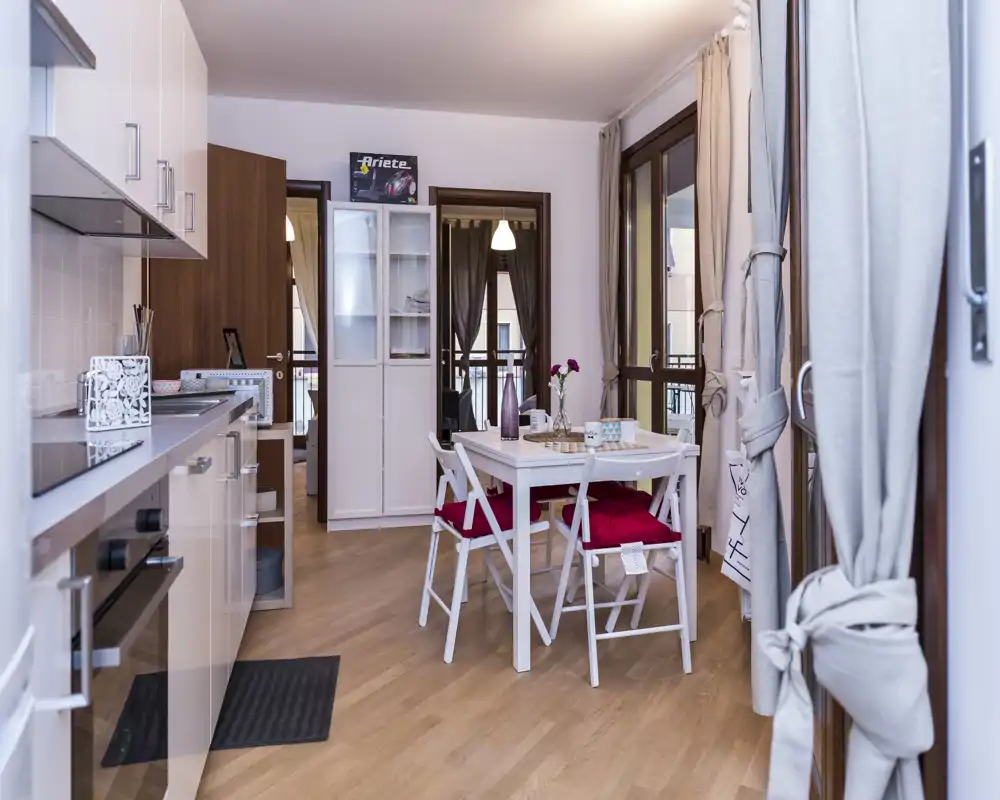 Cucina moderna e luminosa a vista con tavolo da pranzo compatto, mobili bianchi e pavimento effetto legno; allestimento curato con cuscini rossi e dettagli decorativi, perfetta per un appartamento in città.