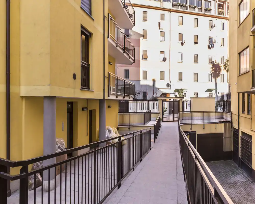 Passerella esterna e cortile comune tra edifici residenziali, con ringhiere, ingressi e terrazze—illuminazione solare del pomeriggio.