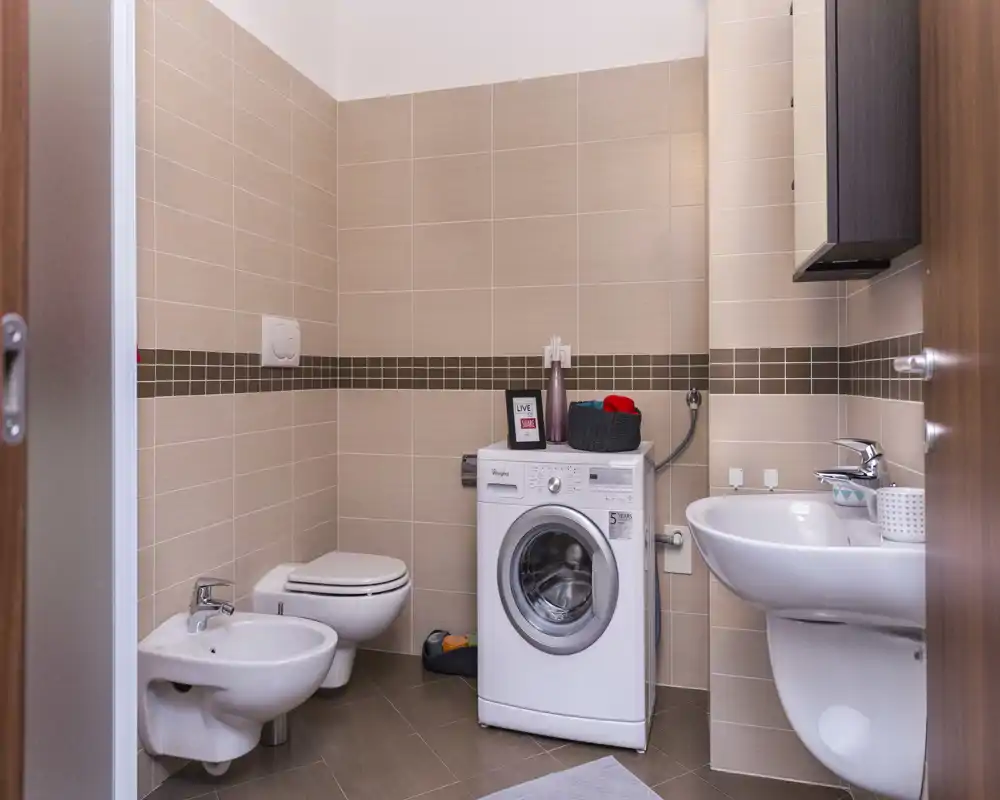 Bagno moderno e luminoso con wc sospeso, bidet, lavabo e lavatrice; piastrelle e colori neutri creano uno spazio pulito e funzionale.