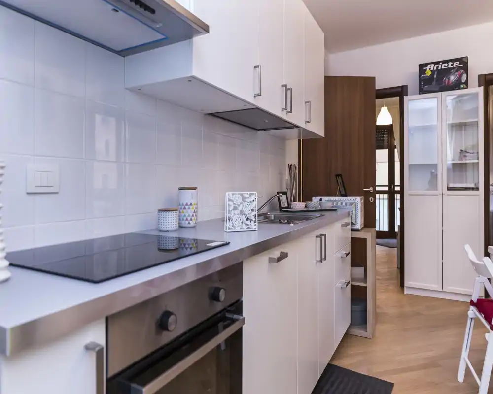 Cucina moderna con piano a induzione, forno incassato e pensili bianchi. Ripiani ordinati, barattoli decorativi e pavimento in legno caldo creano un ambiente cucina curato e accogliente.