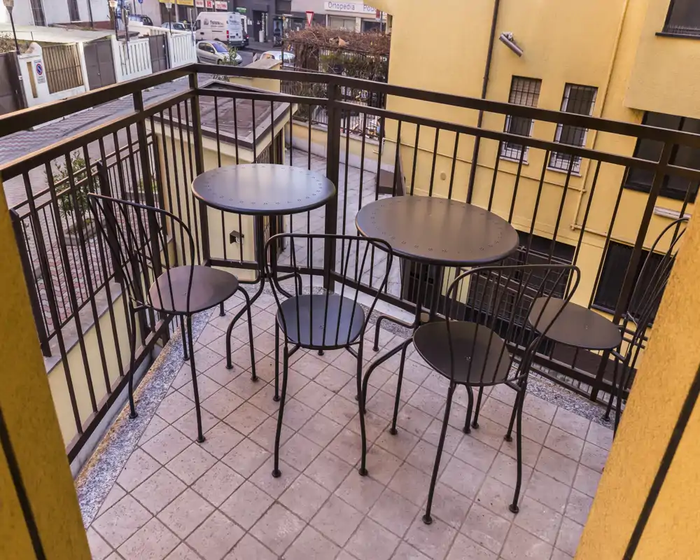 Balcone accogliente con due piccoli tavoli rotondi e quattro sedie in metallo su pavimentazione in piastrelle, protetto da ringhiera metallica — ideale per un caffè mattutino o un pasto leggero all'aperto.