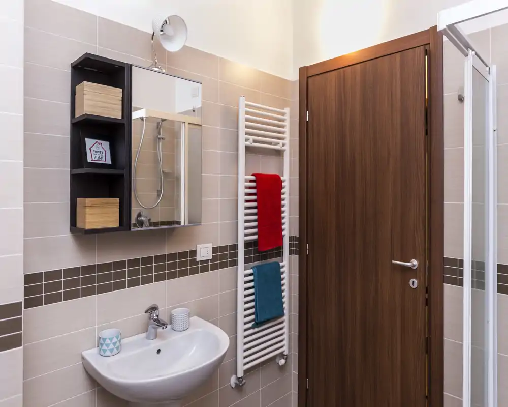 Bagno moderno e pulito con lavabo a parete, mobiletto con specchio, scalda-asciugamani e angolo doccia visibile accanto a una porta in legno.