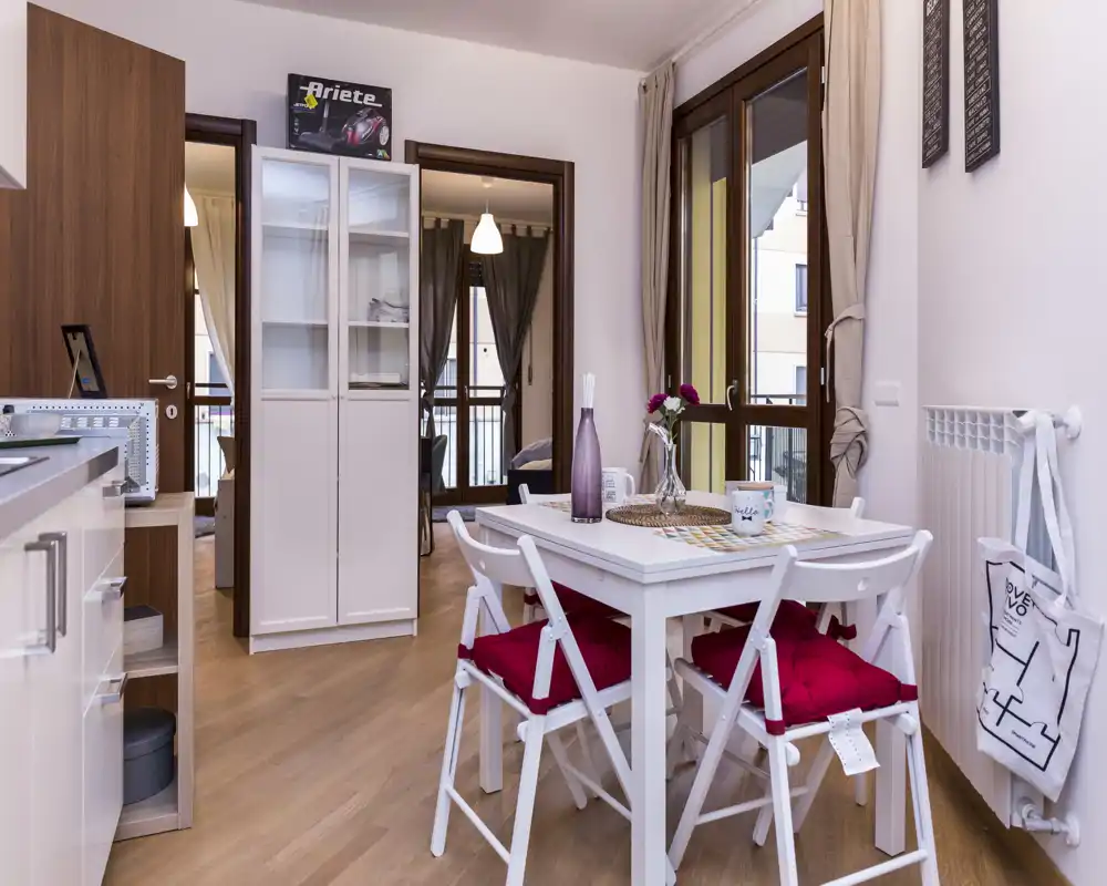 Angolo pranzo moderno e luminoso con tavolo bianco e quattro sedie con cuscini rossi, posizionato vicino a grandi finestre che assicurano luce naturale e un'atmosfera accogliente.