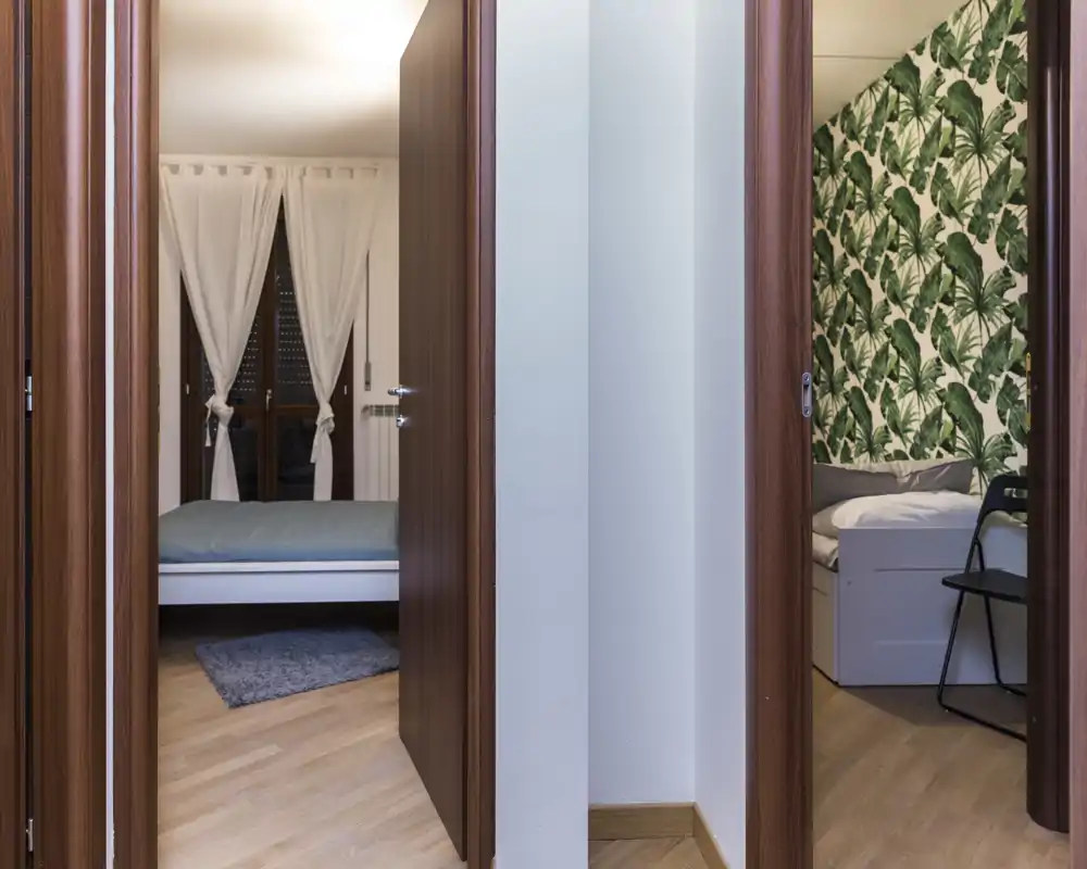 Vista del corridoio con due camere viste tramite le porte aperte: un letto matrimoniale a sinistra con tende morbide e una stanza più piccola a destra con carta da parati tropicale. Pavimenti chiari e porte in legno calde creano un ambiente accogliente.
