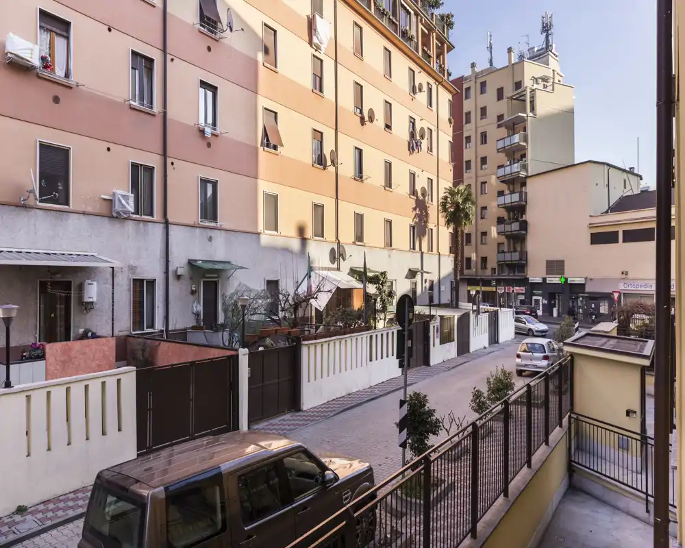 Vista su strada da un balcone: palazzine residenziali, auto parcheggiate e contesto urbano illuminato dalla luce calda del giorno — perfetto per mettere in evidenza posizione e contesto.