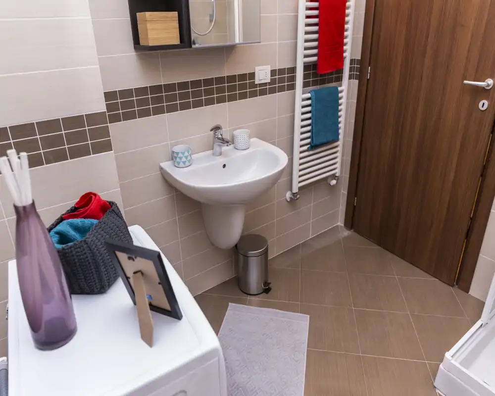 Bagno moderno e pulito con lavabo a parete, scalda-asciugamani e porta in legno; piastrelle neutre ben illuminate e piccoli dettagli decorativi rendono lo spazio ordinato.