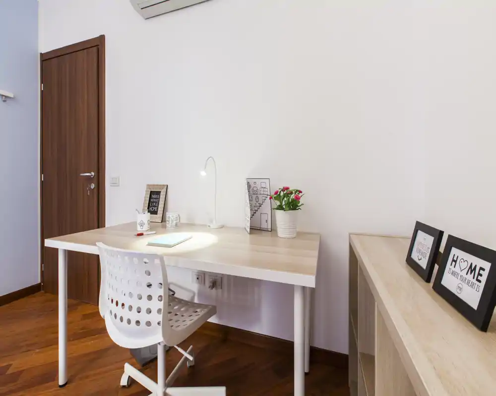 Rincón de trabajo luminoso y minimalista con escritorio de madera clara, silla blanca ergonómica, lámpara de mesa y marcos decorativos — una zona de trabajo ordenada dentro del espacio.