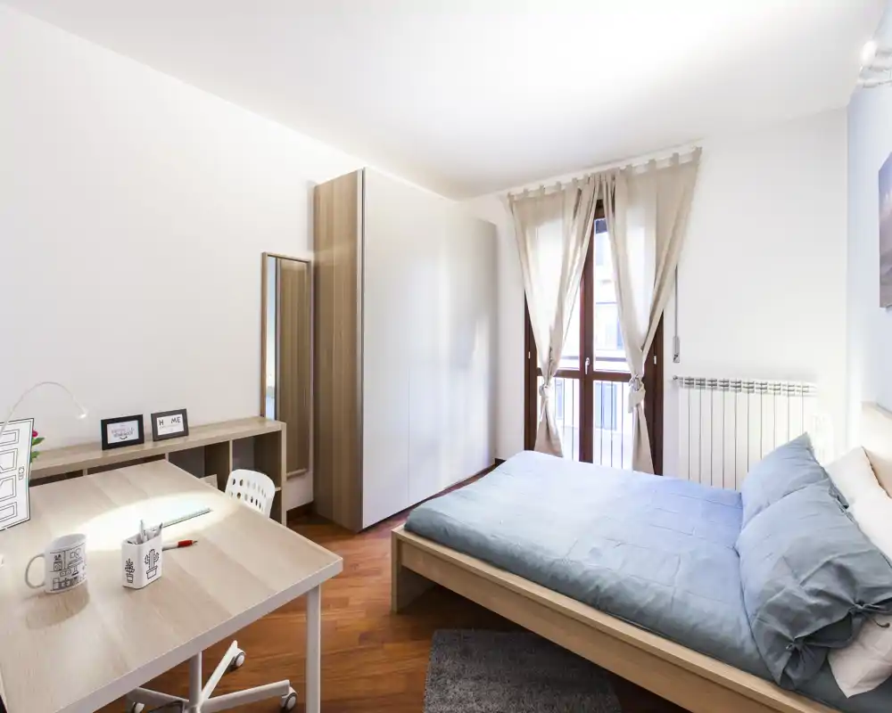 Dormitorio luminoso y ordenado con cama doble, suelo de madera y escritorio junto a la ventana — perfecto para anuncios que destaquen confort y luz natural.
