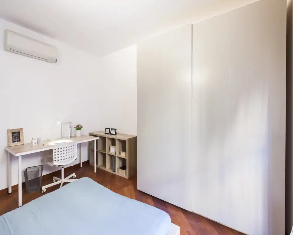 Dormitorio moderno y luminoso con cama doble cubierta con edredón azul claro, zona de trabajo con escritorio y silla, y un gran armario blanco. Líneas limpias, suelo de madera y decoración sencilla crean un espacio tranquilo y funcional, ideal para alquiler.