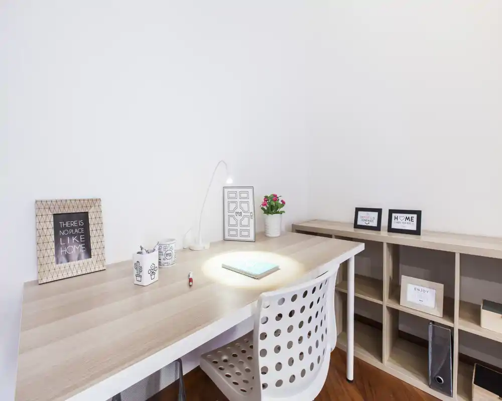 Espacio de trabajo luminoso y minimalista con escritorio de madera clara, silla moderna perforada, lámpara pequeña y marcos decorativos — un rincón ordenado ideal para teletrabajo o estudio.