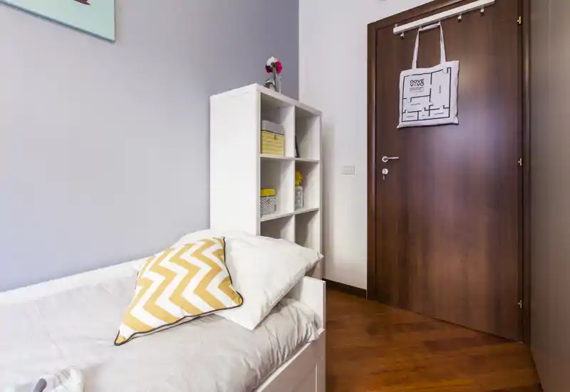 Bright, tidy single bedroom with a visible bed, decorative chevron cushion and open shelving—well lit and neatly staged for rental photos.